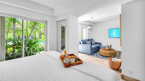 Habitació d'hotel moderna amb un llit gran, una zona de dinar amb un plat de desigini, un sofà blau i una finestra gran amb vista a un paisatge tropical.