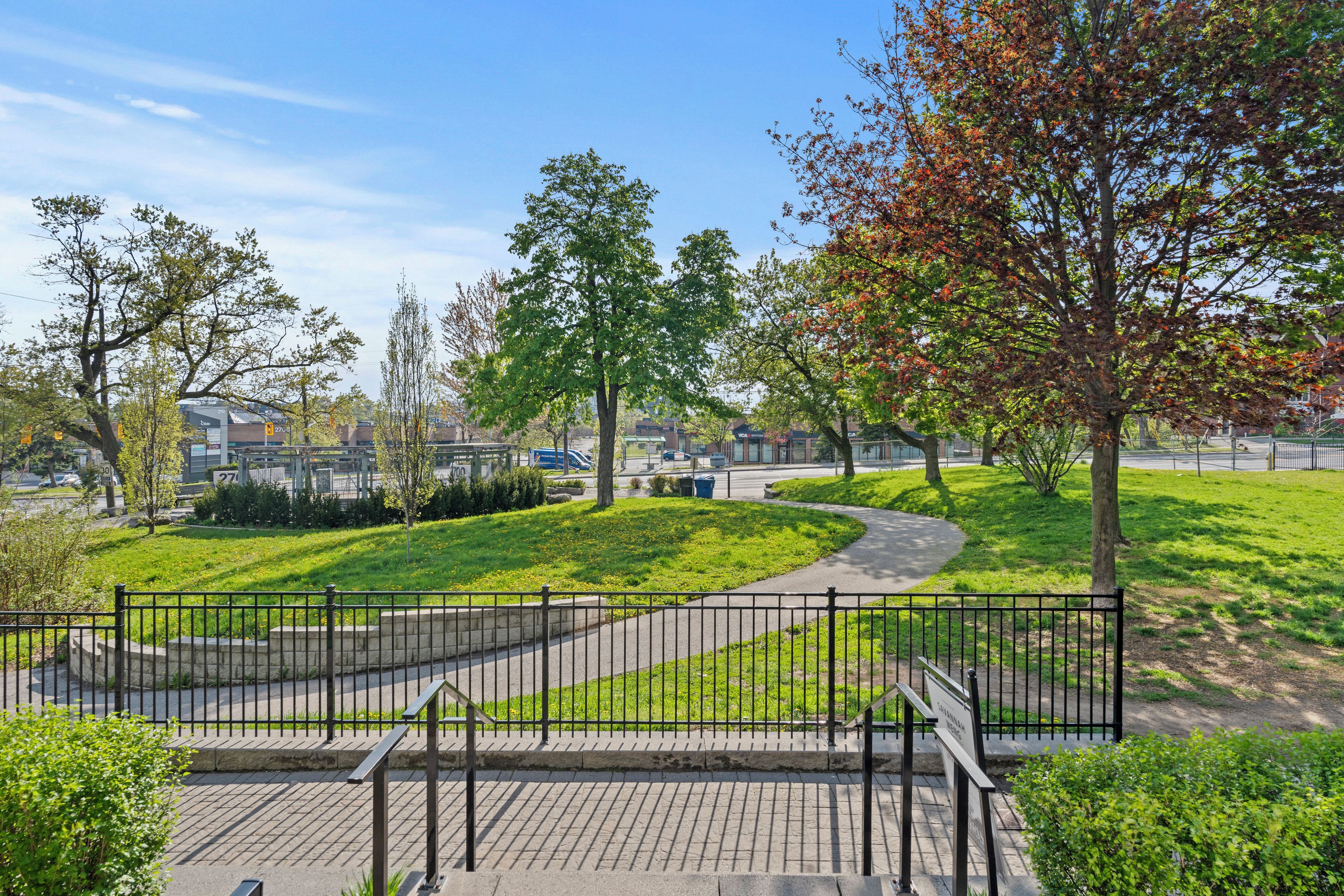 A paved walkway with a metal fence, surrounded by grass and trees.