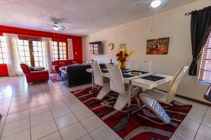 A dining area with a white table, chairs, and a red rug. There is a red sofa and a TV in the background.