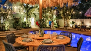 An outdoor dining area with a pool, surrounded by tropical plants and a thatched roof. - Walk to Beach / Shared Heated Pool / LostShaker Two (Lauderdale-by-the-Sea)