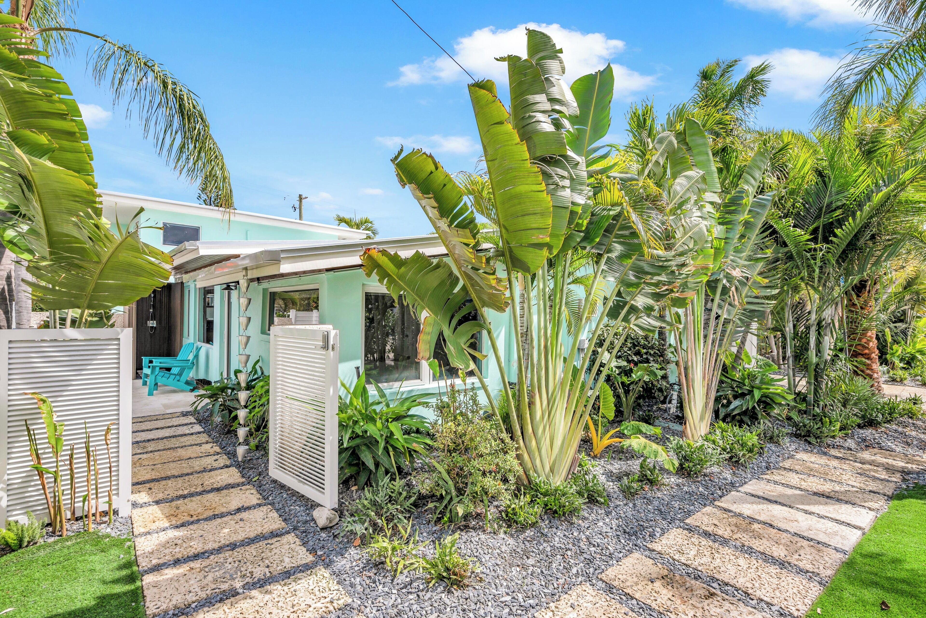A tropical house with a pathway leading to a covered seating area, surrounded by lush greenery and large banana plants.