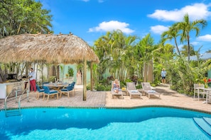 A swimming pool with lounge chairs, a thatched-roof structure, and surrounding palm trees. - Walk to Beach / Shared Heated Pool / Lost Shaker Seven (Lauderdale-by-the-Sea)