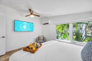 A bedroom with a bed, a ceiling fan, a mounted TV, a window with a view of greenery, and a tray with breakfast items. - Walk to Beach / Shared Heated Pool / Lost Shaker Eight (Lauderdale-by-the-Sea)