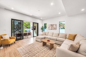 Una sala de estar moderna con un sofá seccional, un sillón amarillo, una mesa de centro y un ventanal de vidrio que da a un espacio exterior.