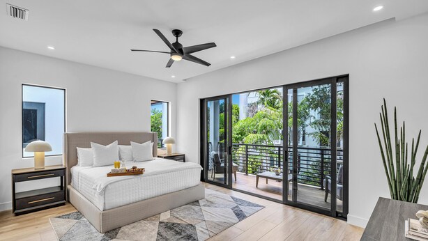 A modern bedroom with a large bed, a ceiling fan, a nightstand with a lamp, and a view of a balcony with outdoor furniture.