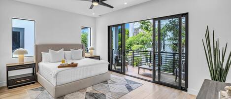 A modern bedroom with a large bed, a ceiling fan, a nightstand with a lamp, and a view of a balcony with outdoor furniture.