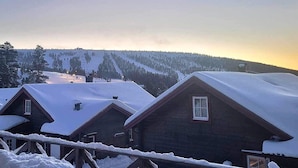 A row of wooden cabins with snow-covered roofs, set against a backdrop of a snowy mountain and a clear sky.