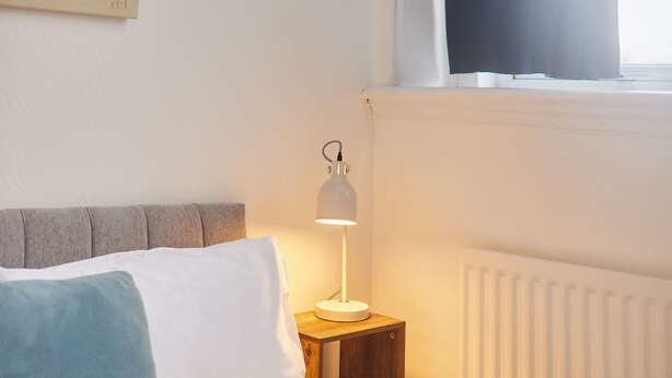 Una cama con almohadillas blancas y azules, una mesita de noche con una lámpara blanca y un radiador.