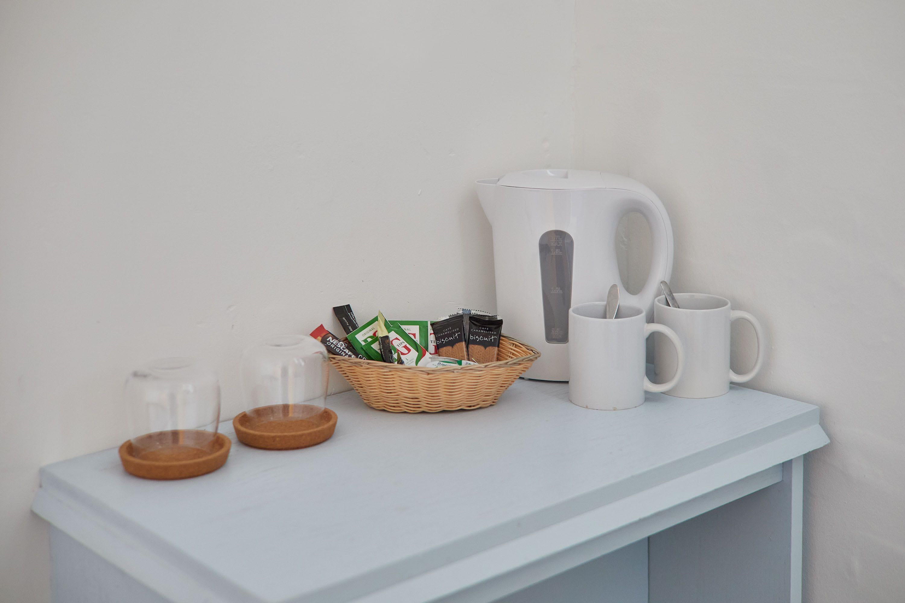 A white kettle, two white mugs, a wicker basket with packets, and two glass containers on a white surface.
