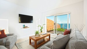 A modern living room with a flat-screen TV, grey sofas, a wooden coffee table, and a large sliding glass door leading to a balcony with a view of the sea.