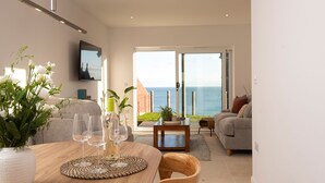 A modern living room with a dining table, chairs, and a view of the sea through large glass doors.