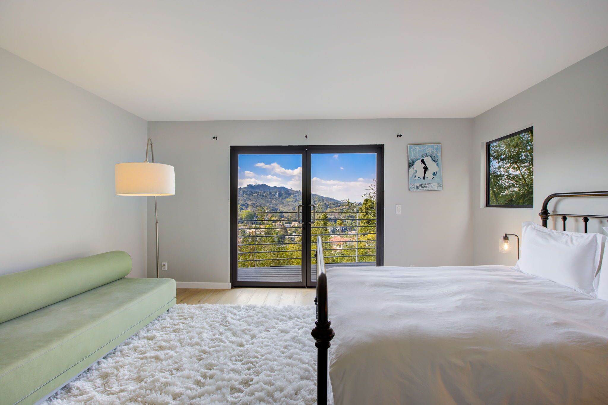 Une chambre avec un lit, un canapé vert, un tapis blanc, une lampe sur pied et une vue sur les montagnes par une grande fenêtre.