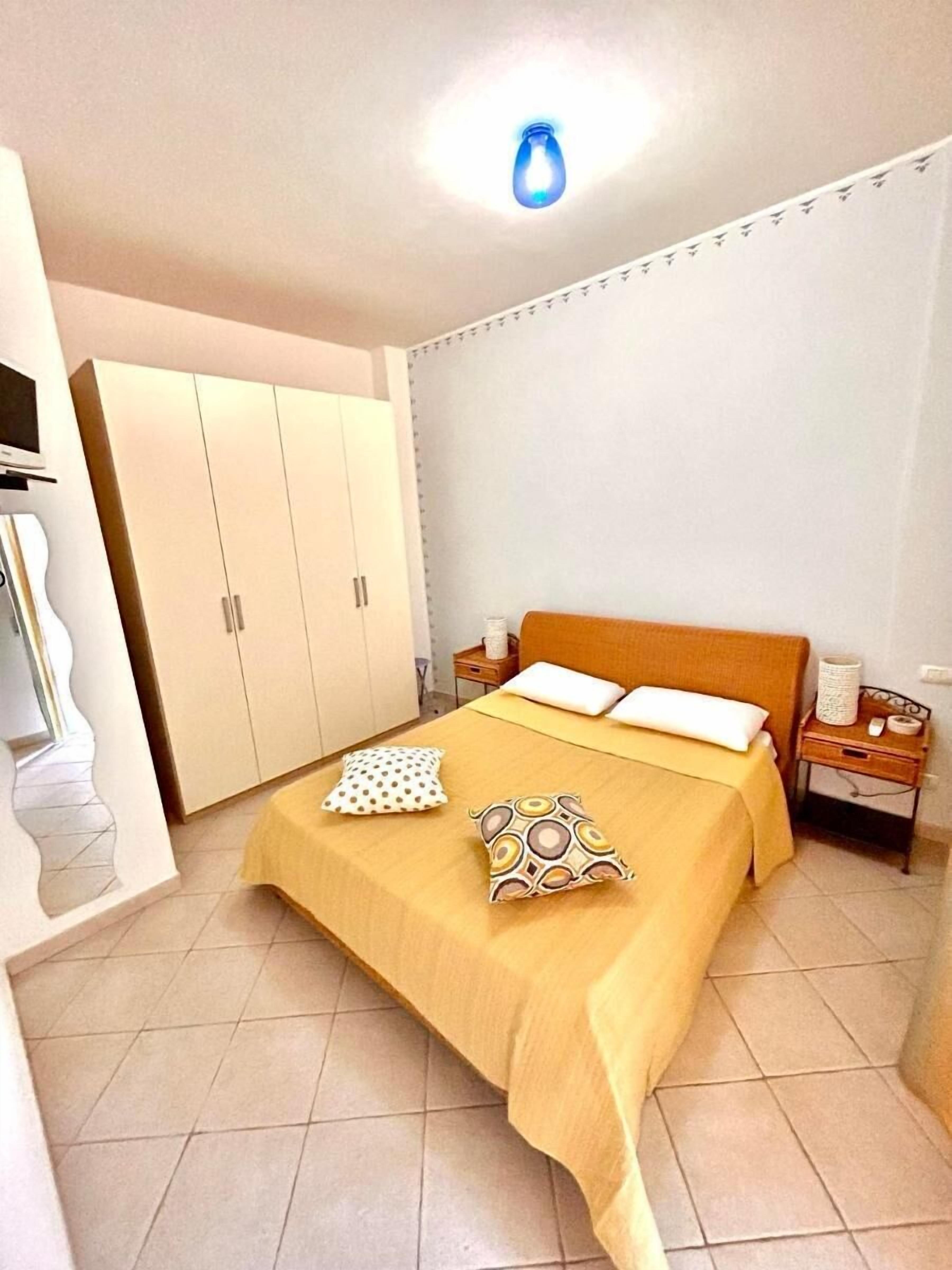 Un dormitorio con cama, dos almohadas, una mesita de noche, un armario y una lámpara en el techo.