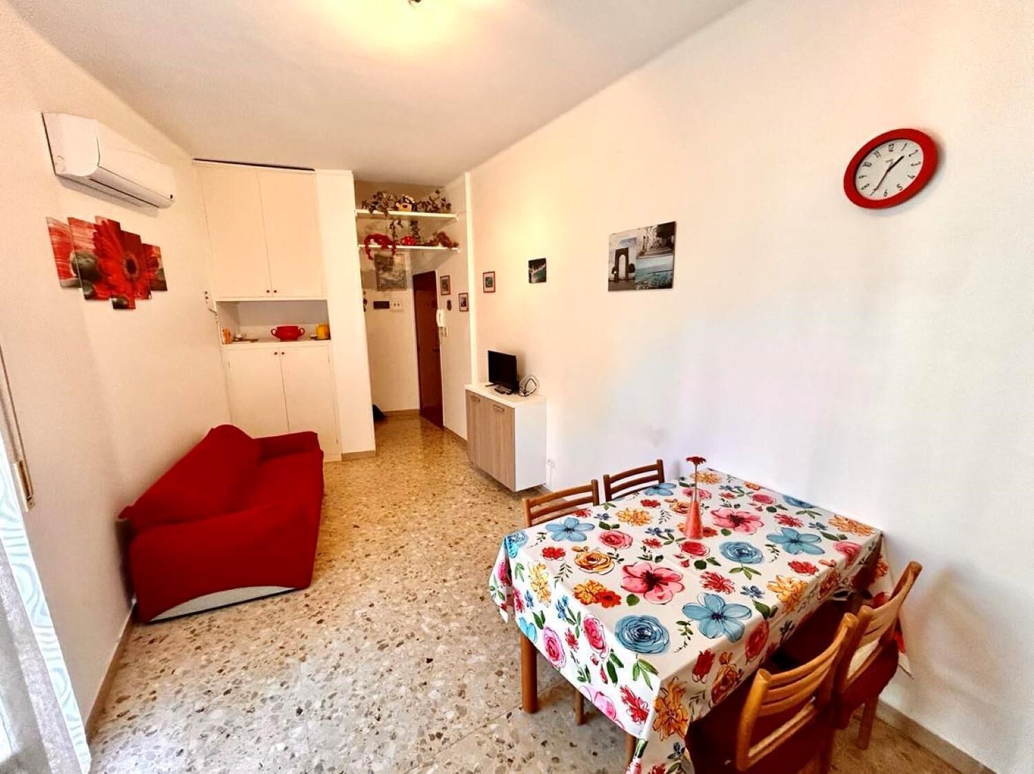 Un espace de vie compact comprenant un coin cuisine, un coin repas avec une nappe à motifs floraux, un canapé rouge et une horloge au mur.