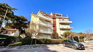 A multi-story residential building with balconies, a parking area, and a clear blue sky. - TRILO EUGENIO MARCELLI - Comfortable three-room apartment with a/c near the sea (Marcelli)