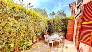 Un patio con tavolo e sedie, circondato da un siepe e un edificio con persiane rosse.