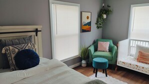 Un dormitorio con una cama, un sillón verde, un taburete pequeño, una cuna y una planta en la pared.