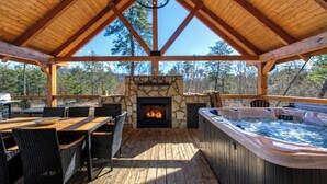 A covered outdoor area with a stone fireplace, a hot tub, and a dining table with chairs. - Makin' Memories Cabin | Hot Tub, Views, Loft, Fire Pit & Game Area + FREE  Tickets (Sevierville)