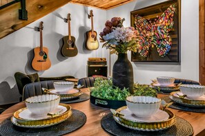 Ein Essbereich mit einem Holztisch, gedeckt für vier Personen, umgeben von Stühlen. An der Wand hängen Gitarren, ein großes gerahmtes Kunstwerk und auf dem Tisch steht eine Vase mit Blumen.