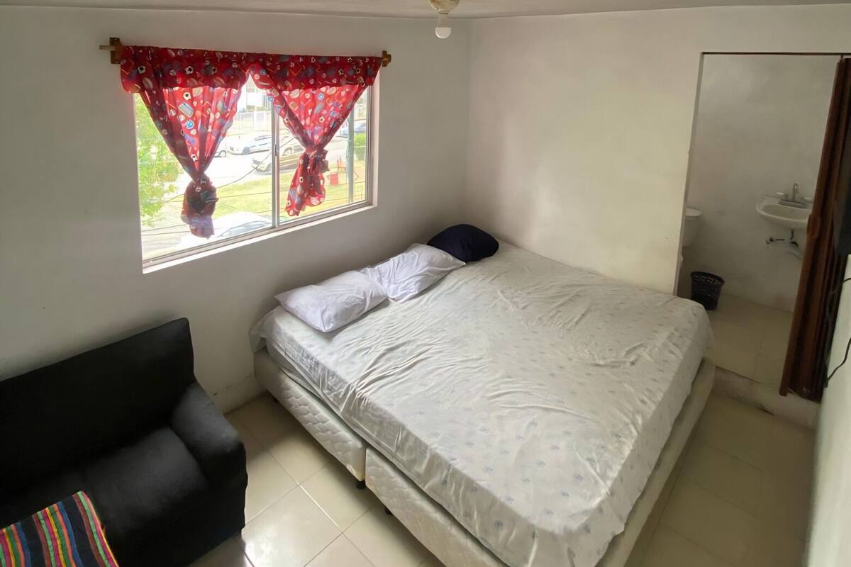 Een kleine kamer met een enkel bed, een raam, een slaapbank en een wastafel.