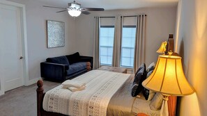 A bedroom with a bed, a desk lamp, a ceiling fan, a window with curtains, a sofa, and a door. - 1120-Spacious 4BR All Suites - 20 min to Disney (Kissimmee)