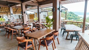 Un restaurante con mesas y sillas de madera, techo de paja y vistas del paisaje.