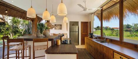 A kitchen with a central island, pendant lights, a ceiling fan, and large windows overlooking a garden.
