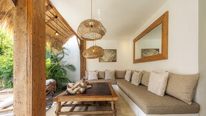 Ein gemütlicher Wohnbereich mit Holztisch, einer Couch mit Kissen, einer hängenden Korbtasche als Leuchte und einem großen Spiegel an der Wand.