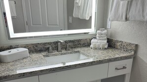 Een badkamer met een granieten aanrechtblad, een witte wastafel en een spiegel erboven. Handdoeken hangen aan de muur en er is een klein schapje met toiletartikelen.
