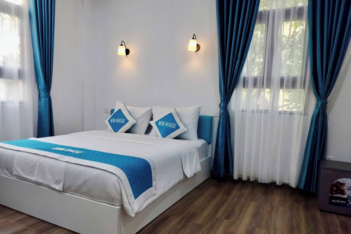 A neatly made bed with a blue accent, two wall-mounted lights, and a window with blue curtains.