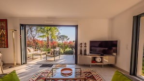 A living room with a glass coffee table, sofa, TV, and a view of a garden through a sliding door. - SAINTE MAXIME Apartment with garden in residence with pool (STE MAXIME)
