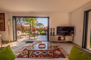 A living room with a glass coffee table, sofa, TV, and a view of a garden through a sliding door. - SAINTE MAXIME Apartment with garden in residence with pool (STE MAXIME)