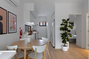 A modern kitchen with white cabinetry, a dining area with a wooden table and white chairs, and a large potted plant. - 2BR Guest House | Pet-Friendly & Peaceful (Niagara Park)