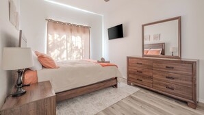 A bedroom with a bed, bedside table, nightstand, dresser, mirror, ceiling fan, and a window with curtains.