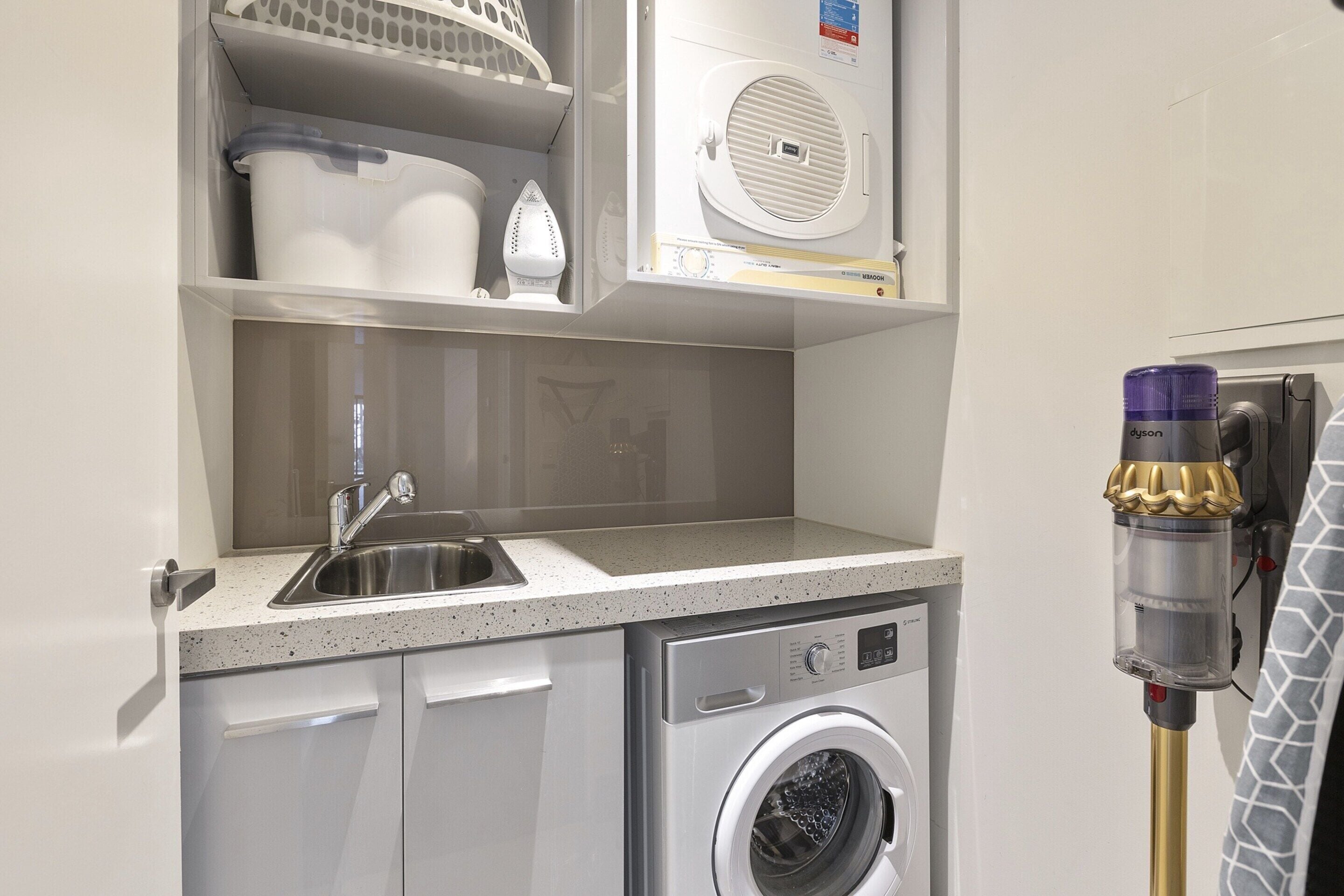 A compact laundry room with a washing machine, a sink, and a Dyson vacuum cleaner.