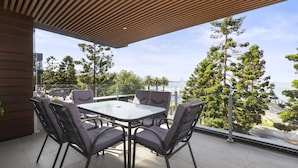 A modern outdoor patio with a glass table and chairs, overlooking a scenic view of trees and a body of water. - Waterfront Apartment With Stunning Bay Views (Geelong)