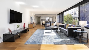 A modern living room with a flat-screen TV, a sofa set, a coffee table, and a large window offering a view of the outdoors. - Waterfront Apartment With Stunning Bay Views (Geelong)