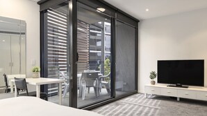 A modern hotel room with a large bed, a TV, a dining area with chairs and a table, and a sliding glass door leading to an outdoor patio. - Waterfront Apartment With Stunning Bay Views (Geelong)