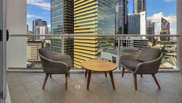 Un balcón con dos sillas de mimbre y una mesita de madera, con vistas a un paisaje urbano de edificios modernos.