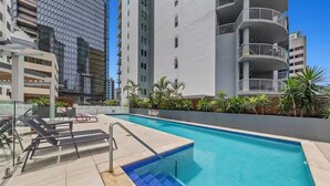 Una piscina con solárium de baldosas, rodeada de edificios modernos y palmeras.