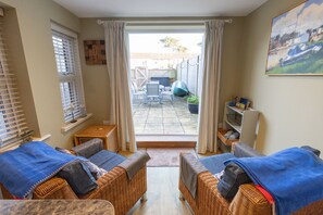A room with wicker furniture, a view to the outside, a small table, and a shelf with items. - Anchor Point - a two bedroom, two bathroom cottage close to Fisherman's Bank, Mudeford (Christchurch)