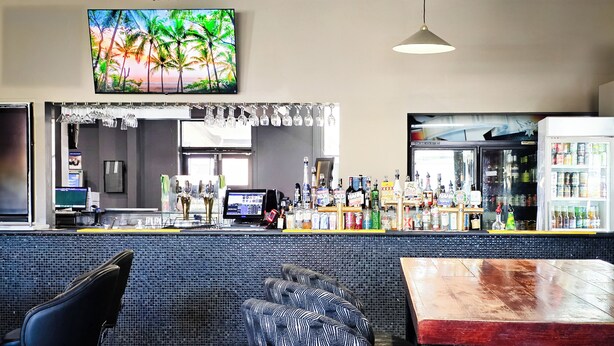 Un espace bar comprenant un comptoir, des tabourets de bar, un téléviseur diffusant des images d’un paysage tropical, ainsi qu’une variété de bouteilles et de verres.