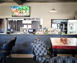 Zona de bar con mostrador, taburetes, televisor mostrando una escena tropical y una variedad de botellas y vasos.