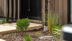 Un edificio moderno con una puerta negra, paneles de madera verticales y un pequeño jardín con plantas y acolchado.