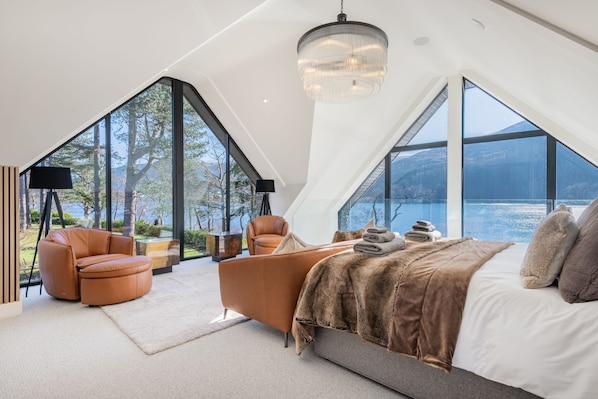 Une chambre moderne avec un grand lit, deux fauteuils en cuir, une cheminée et une vue sur un lac.