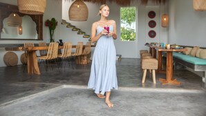 Donna con un vestito azzurro chiaro che tiene un drink in un interno moderno con zona pranzo, posti a sedere e una scala.