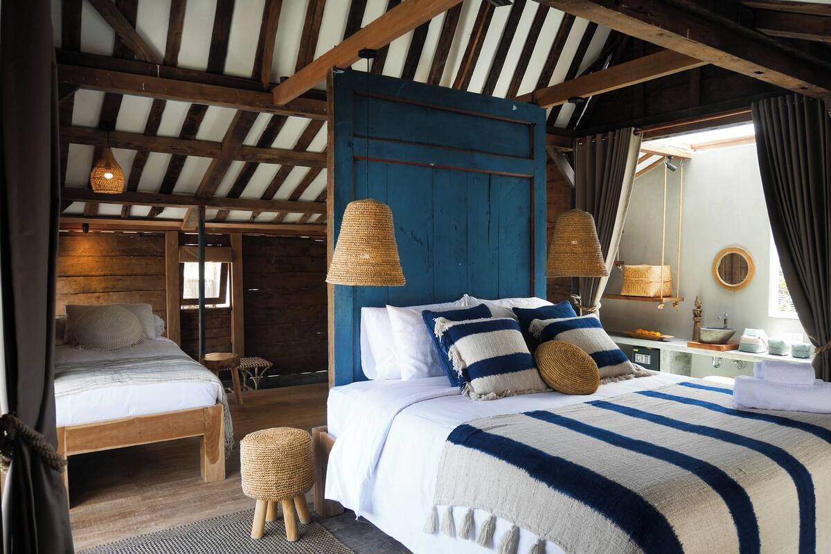 Ein Zimmer mit einem großen Bett, einem kleineren Bett, einer Holzbalkendecke, einer blauen Tür und einem kleinen Tisch.