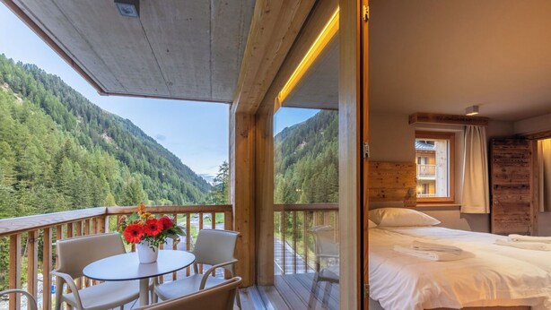 Quarto com cama, mesa e cadeiras, e vista para uma montanha arborizada.