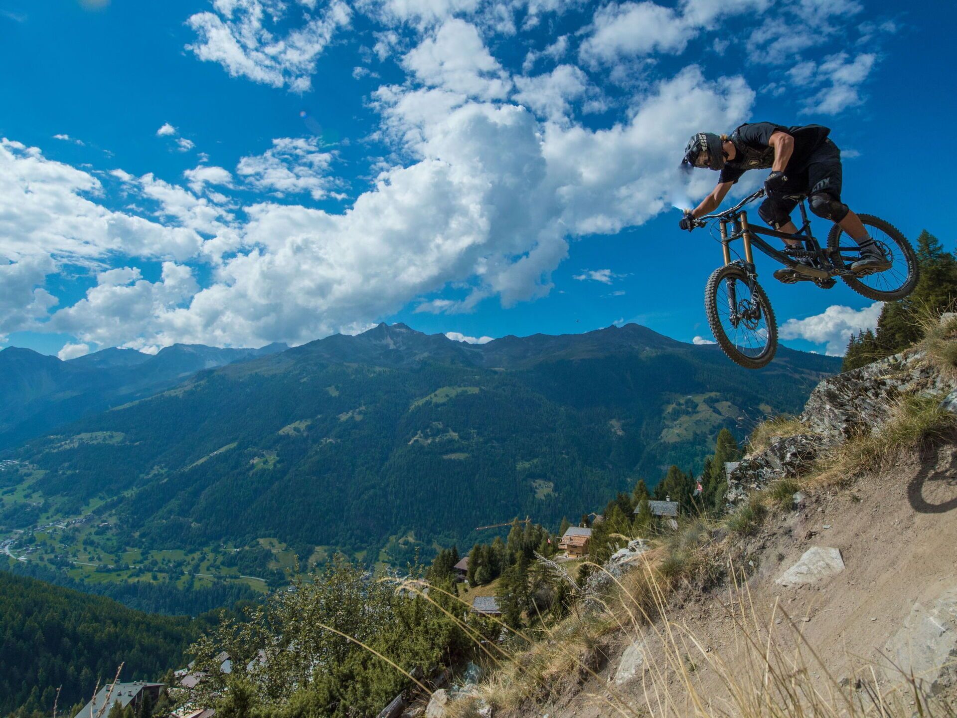 A mountain biker in mid-air on a dirt trail with a mountainous landscape in the background.
