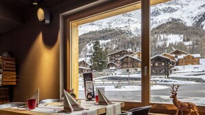A dining area with a table set for two, a wooden interior, and a view of snow-covered buildings and mountains through large windows. - Beautiful apartment for 6 people with WIFI, terrace and pets allowed (Zinal)